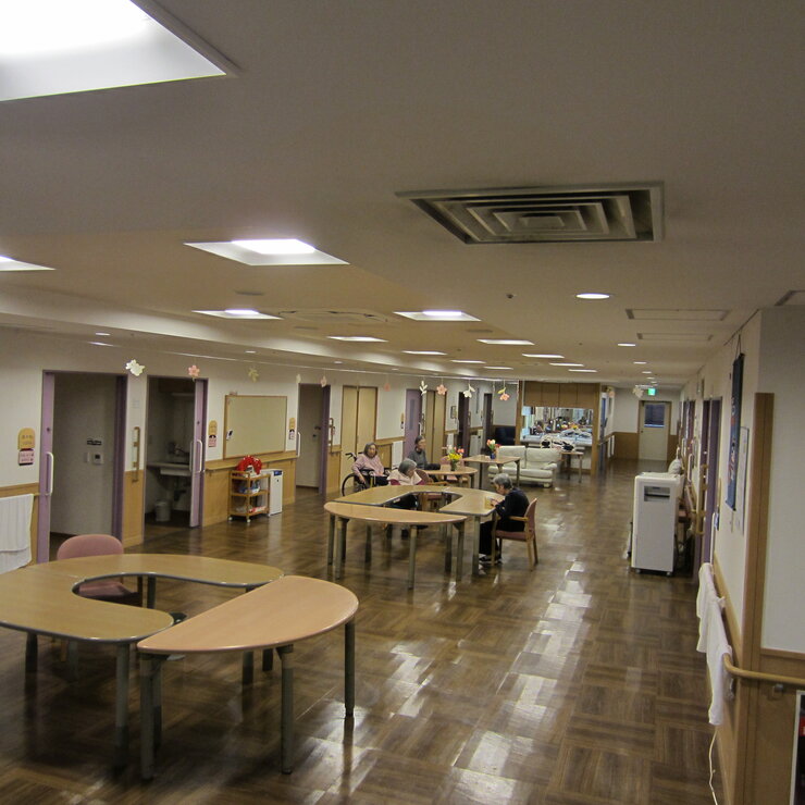 介護施設の共用部。広々とした食堂兼リビングで、複数のテーブルと入居者が見られる。